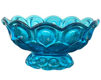 May include: A turquoise glass bowl with a scalloped rim and a pedestal base. The bowl features a star and moon design, and is likely vintage. The bowl is a vibrant shade of blue and would be a decorative piece.