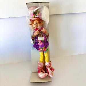 May include: A whimsical decorative elf doll with red hair, round glasses, and a purple velvet coat. The elf wears a plaid shirt, floral pants, yellow socks, and pink shoes. A pink and white hat with floral accents tops the elf's head.