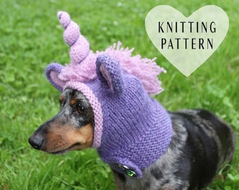 Op de afbeelding: Een teckel die een paarse en roze gebreide eenhoornmuts draagt met een hoorn. De muts heeft een pluche manen en een groene knoop aan de zijkant. De hond staat op groen gras. De tekst "Knitting Pattern" staat in een hartvorm boven de hond.