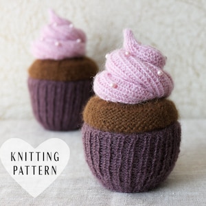 May include: Two knitted cupcake toys with pink frosting and brown cupcake bases. The frosting is decorated with small white beads. The cupcakes are sitting on a white surface. The text "Knitting Pattern" is on a white heart shape in the foreground.