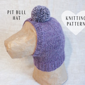 May include: Purple knitted dog hat with a grey pom pom on top. The hat is on a brown mannequin head. The text "PIT BULL HAT" is above the hat and "KNITTING PATTERN" is in a white heart shape to the right of the hat.