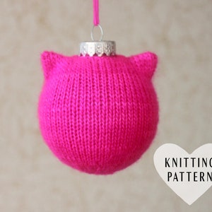 Puede incluir: Un adorno navideño de punto rosa con forma de gato con un colgador de adorno plateado. El adorno tiene orejas de gato en la parte superior.  KNITTING PATTERN está escrito en texto blanco sobre una forma de corazón blanco.