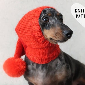 May include: A dachshund wearing a red knitted hat with a pom-pom. The hat has a long neck piece and covers the dog's ears. The dog is looking to the right. The text "KNITTING PATTERN" is in a white heart shape in the upper right corner of the image.