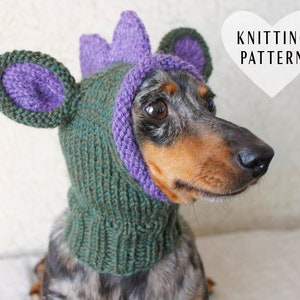 May include: A dachshund dog wearing a green and purple knitted hat with ears and a dinosaur-like spike on top. The hat has a knitted pattern and is designed for pets.
