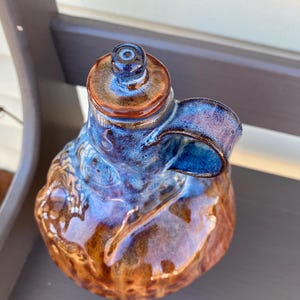 May include: A handmade ceramic bottle with a blue and brown glaze. The bottle has a small handle and a round top.