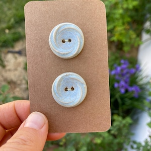 May include: Two light blue ceramic buttons with a swirl design. The buttons have two holes each.
