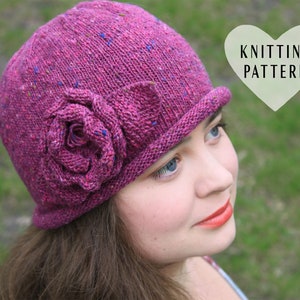 Puede incluir: Un gorro de punto rosa con una gran flor en forma de rosa en el lateral. El gorro tiene un ala ancha y está hecho con un hilo jaspeado. El texto "Knitting Pattern" es visible en un recuadro en forma de corazón.