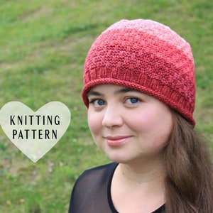 Puede incluir: Una mujer lleva un gorro de punto rojo y rosa con un patrón de color degradado. El gorro está hecho con un punto texturizado. El texto "Knitting Pattern" es visible en forma de corazón.