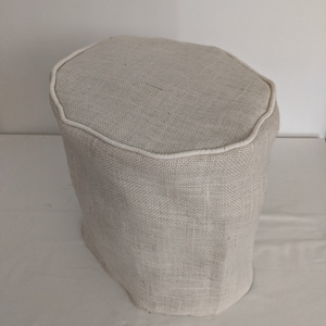 Op de afbeelding: Een beige, achthoekige, stoffen opbergottoman met een witte rand rond de bovenkant.