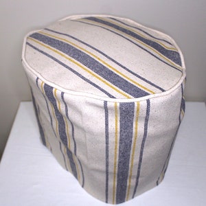Pode incluir: Um item de tecido cilíndrico com uma parte superior arredondada. O tecido é branco sujo com listras verticais em azul marinho e amarelo. A borda superior é aparada com uma borda branca. O item parece ser uma capa para eletrodomésticos de cozinha.