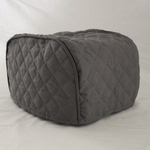 May include: A gray quilted toaster cover. The cover is a rectangular shape with rounded corners and a diamond pattern. The fabric appears to be a soft, durable material. The cover is designed to fit over a standard toaster.