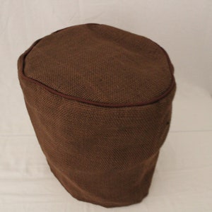 May include: A brown round ottoman cover made of burlap with a brown trim around the top edge.