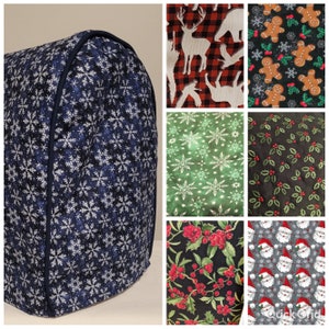 May include: A quilted appliance cover with a navy blue background and white snowflake pattern. The cover is trimmed with a dark blue binding. Additional fabric swatches show festive patterns including reindeer, gingerbread men, and Santa faces.