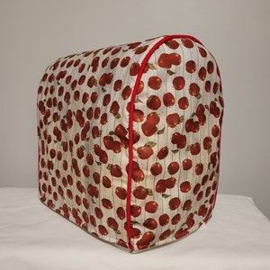 May include: A white and red food processor cover with a repeating pattern of red apples on a white wood background.