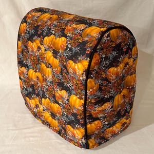 May include: A black and orange kitchen appliance cover with a pumpkin pattern. The cover is made of fabric and has a black trim.