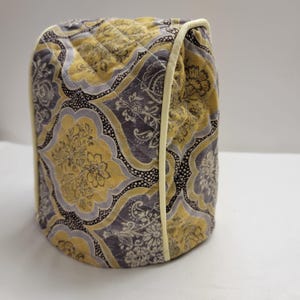 Puede incluir: Una funda acolchada para tostadora con un estampado floral amarillo, gris y blanco. La funda tiene un ribete color crema y un diseño acolchado en forma de diamante. La tela tiene un aspecto de damasco.