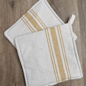 May include: Two square, off-white pot holders with a vertical, golden-yellow stripe design. Each pot holder has a small loop for hanging. The pot holders are made of a textured fabric.