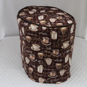 Pode incluir: Capa de cafeteira de tecido castanho com um padrão castanho e branco de chávenas de café e grãos de café. A capa tem uma guarnição castanha em torno do topo.