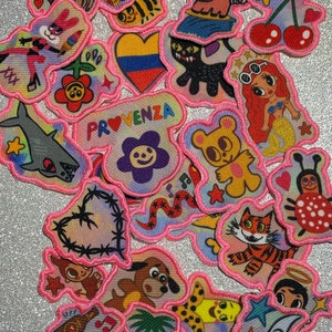 May include: A collection of colorful iron-on patches featuring various cartoon characters and designs. The patches are outlined in pink and have a variety of themes, including animals, food, and nature. Some of the patches include text, such as "Provenza" and "Shroom Queens".