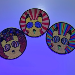 May include: Three round embroidered patches featuring a cartoon character with psychedelic eyes and a green drool. The patches have different backgrounds: red and white stripes, rainbow, and a colorful polka dot pattern.