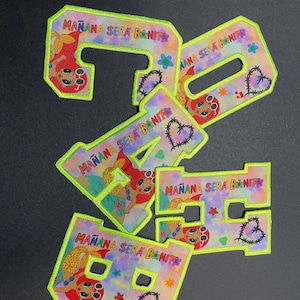 May include: Five neon green embroidered patches with a tie-dye background. Each patch features a cartoon mermaid with red hair and sunglasses. The text "MAÑANA SERA BANIT" is written on each patch.