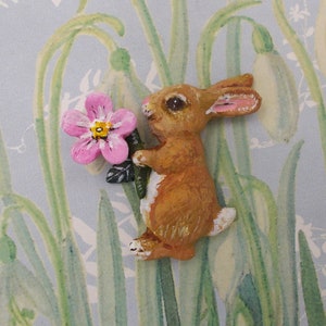 Puede incluir: Una figurita de conejo marrón sosteniendo una flor rosa con un centro amarillo. La figurita está pintada con detalles realistas y se coloca sobre un fondo azul y verde.