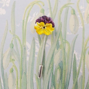 May include: A yellow and purple pansy flower pin with a silver stem. The pin is on a background of a watercolour painting of white flowers.