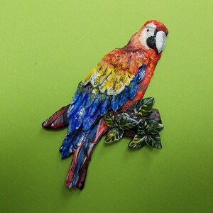 Puede incluir: Una figurita de guacamayo rojo, amarillo y azul con una rama verde frondosa. El guacamayo está posado en la rama con las alas extendidas.