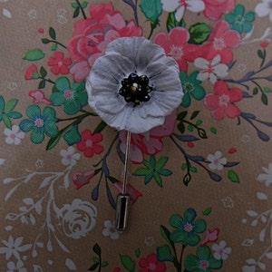 Puede incluir: Broche de flor blanca de fieltro con un centro negro, fijada a un fondo de tela con estampado floral.