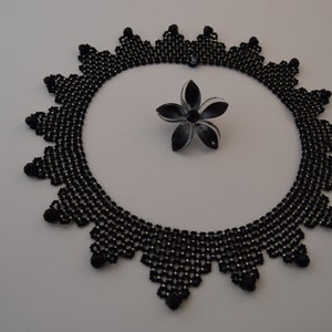 May include: A black beaded necklace with a delicate, intricate design. The necklace is made of small, round beads that are arranged in a pattern of overlapping triangles. The necklace is circular and has a diameter of approximately 10 inches.