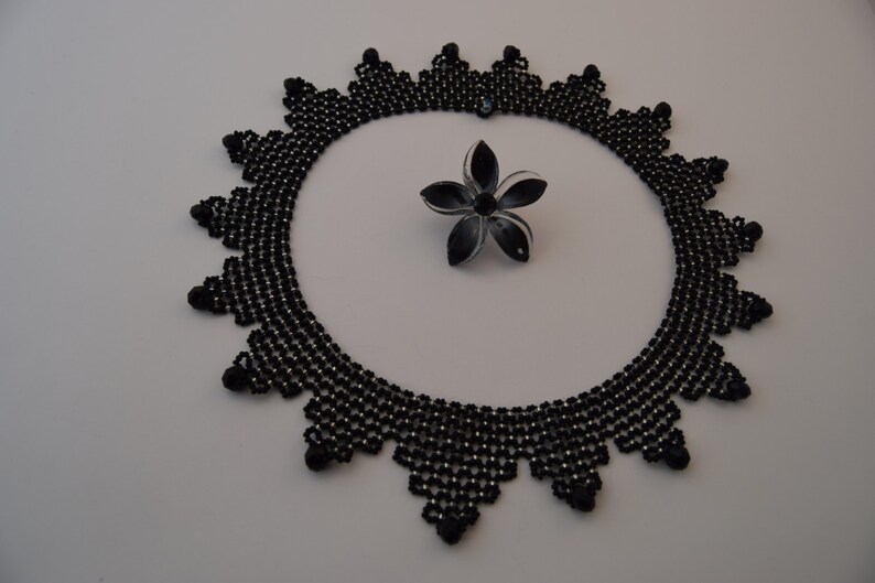 May include: A black beaded necklace with a delicate, intricate design. The necklace is made of small, round beads that are arranged in a pattern of overlapping triangles. The necklace is circular and has a diameter of approximately 10 inches.