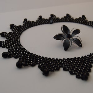 May include: A black beaded necklace with a delicate, intricate design. The necklace is laid out on a white surface with a black flower-shaped brooch.