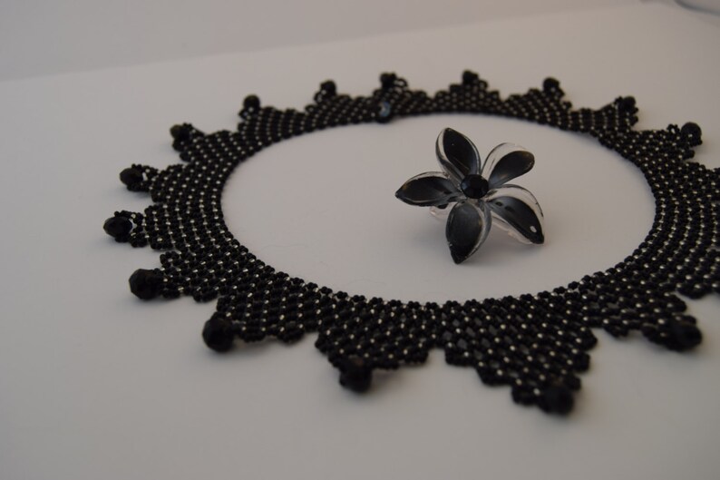May include: A black beaded necklace with a delicate, intricate design. The necklace is laid out on a white surface with a black flower-shaped brooch.