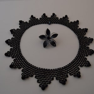 May include: A black beaded necklace with a geometric design. The necklace is made of small black beads and has a scalloped edge. A black and white flower brooch is in the center of the necklace.