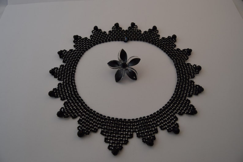 May include: A black beaded necklace with a geometric design. The necklace is made of small black beads and has a scalloped edge. A black and white flower brooch is in the center of the necklace.