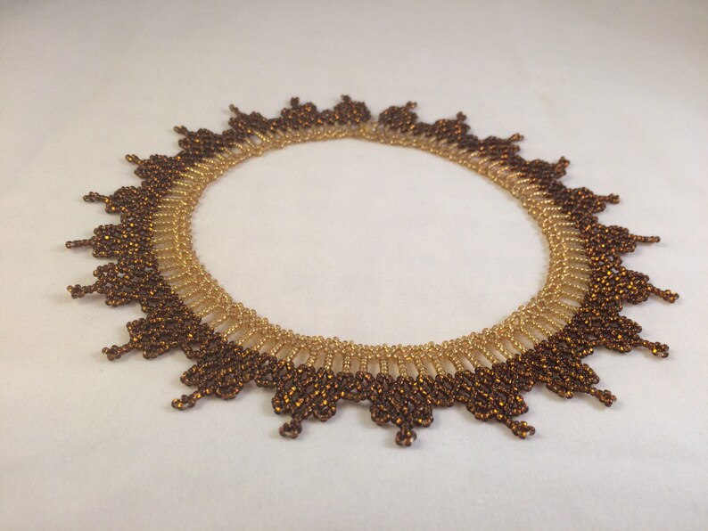 May include: A brown and gold beaded necklace with a scalloped edge. The necklace is made of small, round beads that are arranged in a pattern. The necklace is a statement piece that would add a touch of elegance to any outfit.