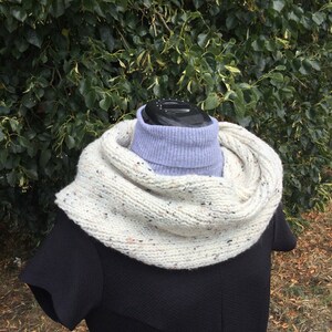 May include: A white and beige cowl neck scarf with a speckled pattern. The scarf is worn over a black mock neck shirt.