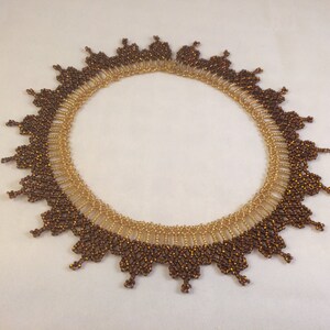 May include: A brown and gold beaded necklace with a scalloped edge. The necklace is made of small, round beads that are arranged in a pattern.