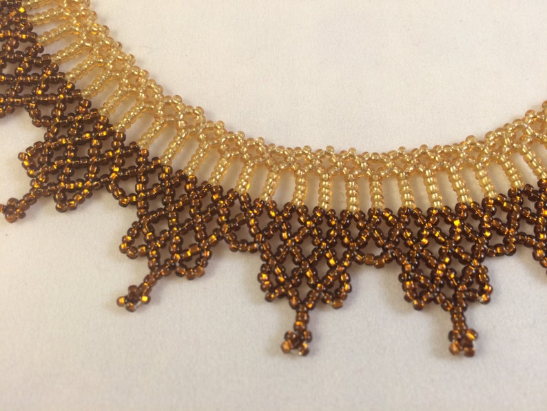 May include: A beaded necklace with a brown and gold color gradient. The necklace is made of small, round beads that are woven together in a delicate pattern.