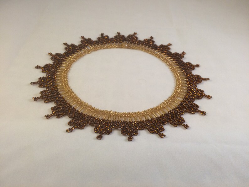 May include: A brown and gold beaded necklace with a scalloped edge. The necklace is made of small, round beads that are arranged in a circular pattern.