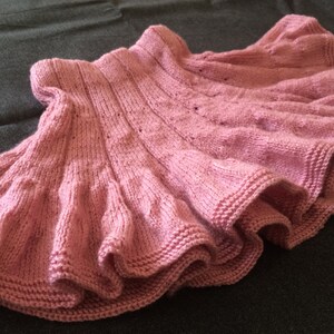 Op de afbeelding: Een roze gebreide rok met een gerimpelde zoom. De rok is gemaakt met een delicaat steekpatroon en heeft een zachte, vrouwelijke uitstraling.