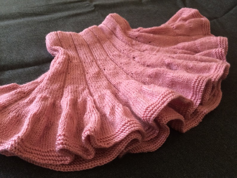 Op de afbeelding: Een roze gebreide rok met een gerimpelde zoom. De rok is gemaakt met een delicaat steekpatroon en heeft een zachte, vrouwelijke uitstraling.