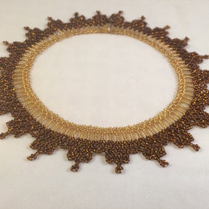 May include: A gold and brown beaded necklace with a scalloped edge. The necklace is made of small, round beads that are arranged in a pattern to create a delicate and intricate design.