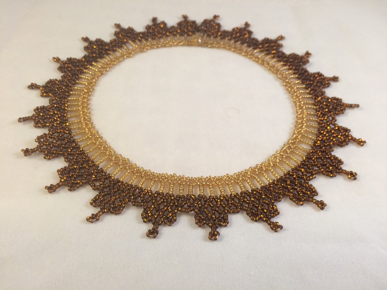 May include: A gold and brown beaded necklace with a scalloped edge. The necklace is made of small, round beads that are arranged in a pattern to create a delicate and intricate design.