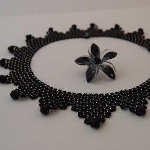 May include: A black beaded necklace with a delicate, intricate design. The necklace is circular and has a scalloped edge. A small black flower is lying next to the necklace.