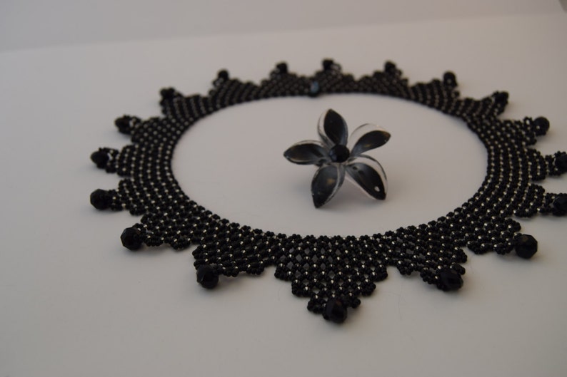 May include: A black beaded necklace with a delicate, intricate design. The necklace is circular and has a scalloped edge. A small black flower is lying next to the necklace.