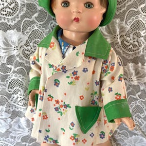 May include: A doll with light brown hair wearing a green bonnet, a cream-colored coat with a multi-colored floral pattern, green cuffs and pockets, a blue collared shirt underneath, white socks, and green shoes with small beige ribbons on the front.