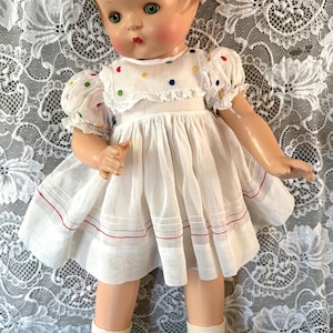 Puede incluir: Una muñeca con cabello rojizo y una diadema verde, vestida con un vestido blanco de lunares coloridos, mangas abullonadas y rayas rojas y azules en el dobladillo. La muñeca lleva calcetines blancos y zapatos verdes con cordones claros.