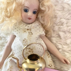 Puede incluir: Una muñeca de porcelana con cabello rubio rizado que lleva un vestido blanco con estampado floral marrón se sienta sobre una superficie rosa con una pequeña tetera dorada.