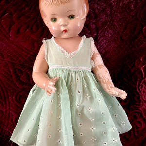 May include: Vintage doll with a painted face, green eyes, and red lips. The doll wears a light green dress with white lace trim and embroidered details. The doll's hair is a reddish-brown color.